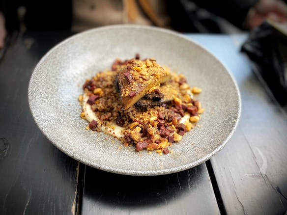 A plate with an elegantly arranged dish featuring what appears to be roasted meat or a similar protein, served over a bed of grains or couscous. The dish is garnished with diced pieces of vegetables or nuts, and is presented on a round, textured plate placed on a dark wooden table.