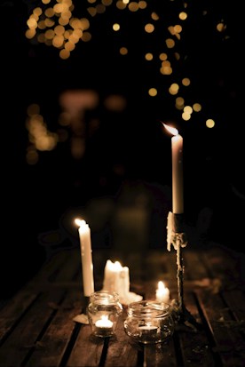 Several candles are arranged on a wooden surface. Some are placed in glass jars, while others are free-standing. The background is dark with a warm bokeh effect from string lights, creating a soft and cozy atmosphere.