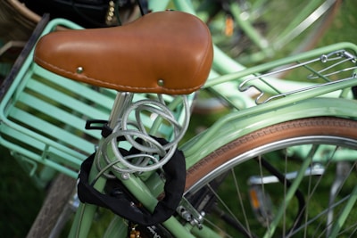 A vintage-style bicycle with a brown leather seat and a green frame. A coiled cable lock is wrapped around the seat post, and the bike features a rear luggage rack. The tire has a tan sidewall, and the background is blurred with another bike partially visible.