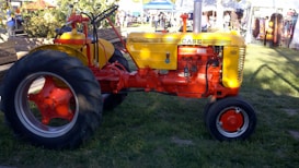 A brightly colored vintage tractor with a yellow hood and red body is parked on a grassy area. The tractor is surrounded by a lively market scene, with people and stalls visible in the background. The tractor’s large rear wheels and smaller front wheels are distinctively red, matching the overall vibrant color scheme.