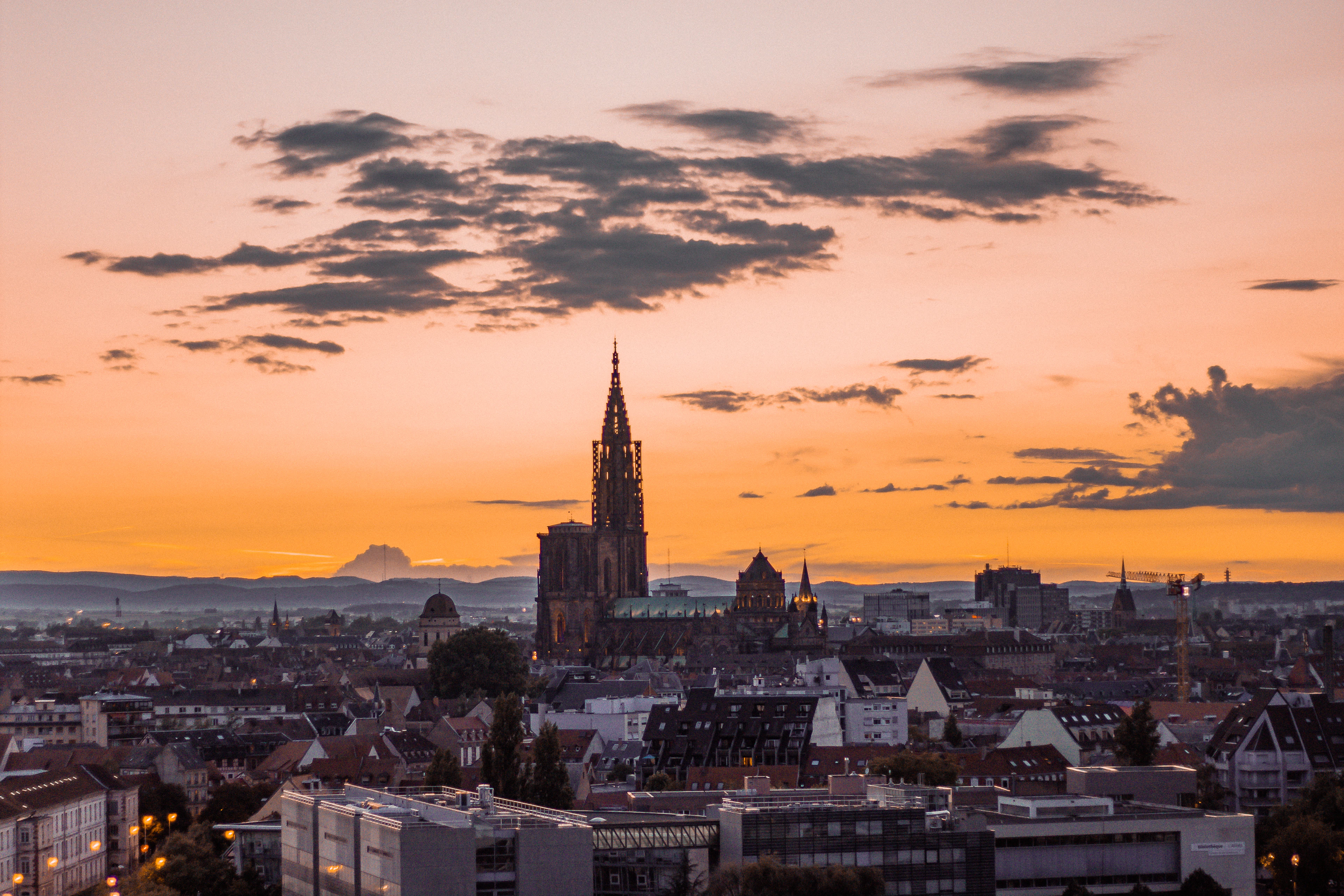 The single spire of Strasbourg Cathedral rising dark above the city's rooftops at sunset, a band of orange sky and drifting cloud behind it and the Vosges hills on the horizon
