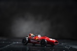 A small red toy race car sits on a dark surface with a blurred smoky background. The car features a sleek design with prominent black wheels and a visible driver's seat.