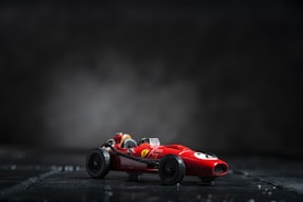 A small red toy race car sits on a dark surface with a blurred smoky background. The car features a sleek design with prominent black wheels and a visible driver's seat.