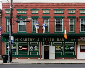 A red brick building features a green facade with the name 'McCarthy's Irish Bar'. It has various neon signs for different beer brands and mannequins dressed in sports outfits on the upper ledge. An Irish flag is displayed prominently.