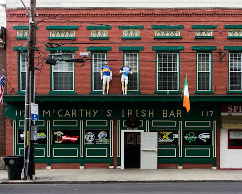 A red brick building features a green facade with the name 'McCarthy's Irish Bar'. It has various neon signs for different beer brands and mannequins dressed in sports outfits on the upper ledge. An Irish flag is displayed prominently.