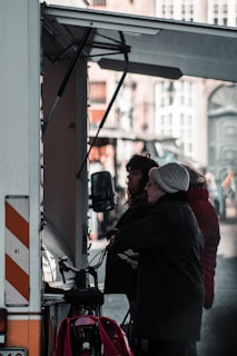 Two people, one of whom appears to be an elderly woman wearing a knit hat and coat, are standing in front of a food truck or market stall. They seem to be making a transaction or interacting with the vendor. A bicycle is parked next to them, and the scene takes place in an urban area with buildings visible in the background.