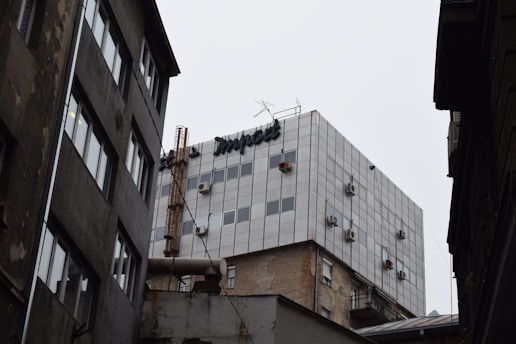 A multi-story building features numerous air conditioning units, with a visible rooftop chimney and antennas. The facade is an array of small, uniformly-sized windows. The surrounding structures appear aged and worn, with contrasting architecture and materials.