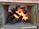 A small furnace containing black charcoal burning with bright orange flames. The surrounding structure is made of cement or fireproof material, indicating a built-in oven or fireplace. The charred wood pieces are consuming the heat, casting an intense glow.