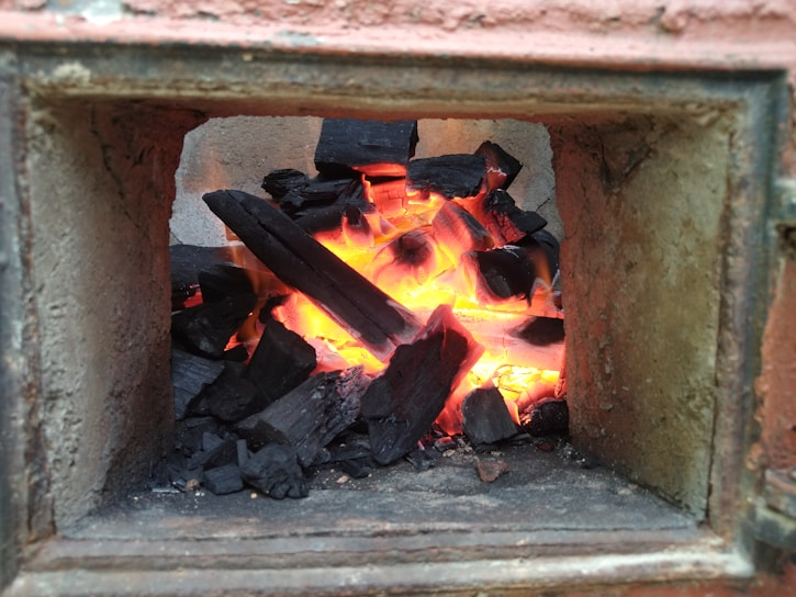A small furnace containing black charcoal burning with bright orange flames. The surrounding structure is made of cement or fireproof material, indicating a built-in oven or fireplace. The charred wood pieces are consuming the heat, casting an intense glow.