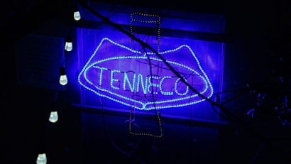 A neon sign with the word 'TENNECO' is illuminated in a bright blue color. It is surrounded by hanging light bulbs, and there are some branches casting shadows over the scene.