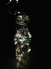 A glass bottle is adorned with small, colorful flower decorations in hues of pink and blue. Inside the bottle, there are tiny, glowing string lights that illuminate brightly, creating a charming contrast against the dark background.