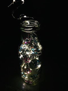 A glass bottle is adorned with small, colorful flower decorations in hues of pink and blue. Inside the bottle, there are tiny, glowing string lights that illuminate brightly, creating a charming contrast against the dark background.