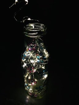 A glass bottle is adorned with small, colorful flower decorations in hues of pink and blue. Inside the bottle, there are tiny, glowing string lights that illuminate brightly, creating a charming contrast against the dark background.