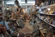 An antique shop filled with a variety of vintage and handcrafted items. Shelves are lined with ceramic vases and bowls, while stacks of intricately patterned rugs and textiles are piled on the floor. Brass and copper pots, along with ornate picture frames and trays, create a sense of cluttered charm. Hanging lamps and artifacts add to the eclectic assortment, illuminated by warm lighting from ceiling fixtures.