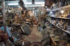 An antique shop filled with a variety of vintage and handcrafted items. Shelves are lined with ceramic vases and bowls, while stacks of intricately patterned rugs and textiles are piled on the floor. Brass and copper pots, along with ornate picture frames and trays, create a sense of cluttered charm. Hanging lamps and artifacts add to the eclectic assortment, illuminated by warm lighting from ceiling fixtures.