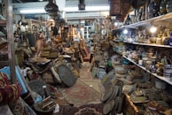 An antique shop filled with a variety of vintage and handcrafted items. Shelves are lined with ceramic vases and bowls, while stacks of intricately patterned rugs and textiles are piled on the floor. Brass and copper pots, along with ornate picture frames and trays, create a sense of cluttered charm. Hanging lamps and artifacts add to the eclectic assortment, illuminated by warm lighting from ceiling fixtures.