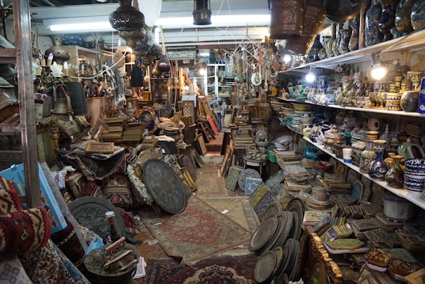 An antique shop filled with a variety of vintage and handcrafted items. Shelves are lined with ceramic vases and bowls, while stacks of intricately patterned rugs and textiles are piled on the floor. Brass and copper pots, along with ornate picture frames and trays, create a sense of cluttered charm. Hanging lamps and artifacts add to the eclectic assortment, illuminated by warm lighting from ceiling fixtures.