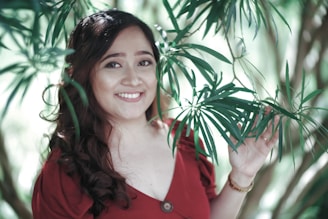 A woman is smiling while standing among green foliage, with long, dark hair cascading over her shoulders. She is wearing a red dress with a deep neckline and holds a branch of leaves.