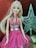 A doll with long blonde hair is dressed in a glittery pink halter-neck dress with a silver belt. The background features a textured red material and a sparkling silver star.