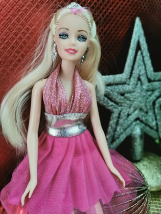 A doll with long blonde hair is dressed in a glittery pink halter-neck dress with a silver belt. The background features a textured red material and a sparkling silver star.