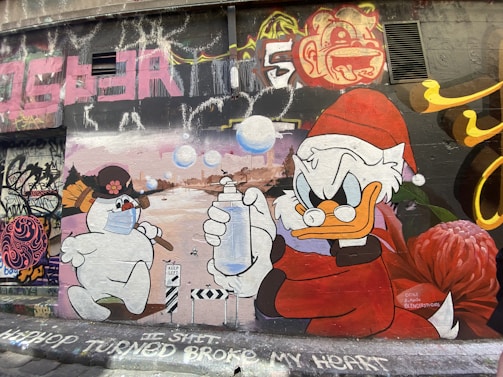 A vibrant street art mural features a cartoon snowman wearing a face mask and holding a broom, facing a duck character dressed in a red Santa outfit holding a spray bottle. The background includes graffiti in various colors, and the words 'HIP HOP TURNED BROKE MY HEART' are written at the bottom.