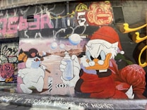 A vibrant street art mural features a cartoon snowman wearing a face mask and holding a broom, facing a duck character dressed in a red Santa outfit holding a spray bottle. The background includes graffiti in various colors, and the words 'HIP HOP TURNED BROKE MY HEART' are written at the bottom.