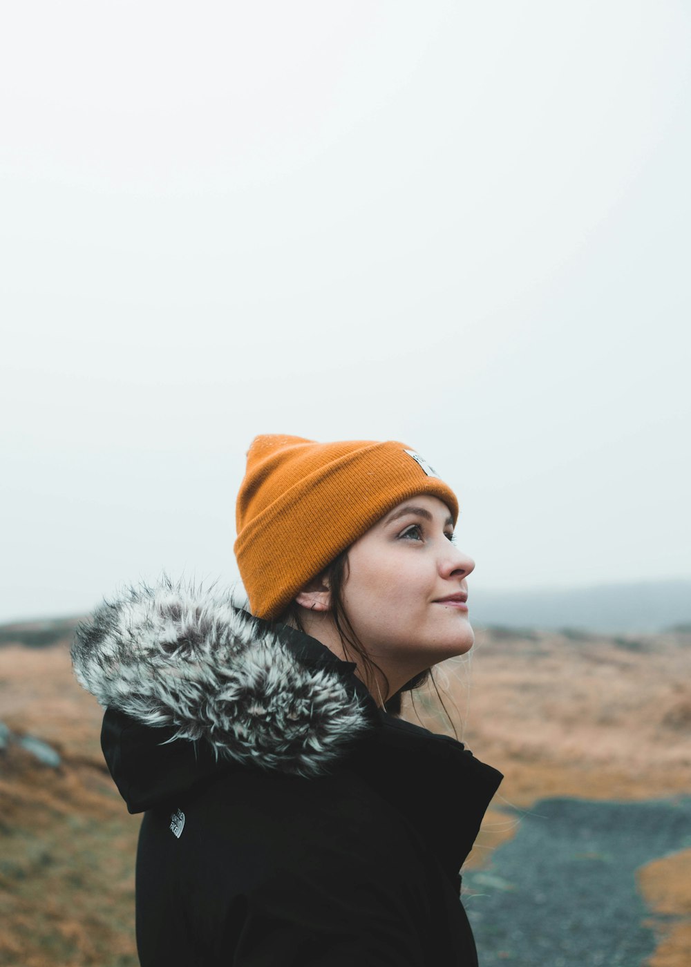 woman in black fur coat and orange knit cap standing on brown sand during daytime