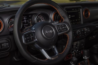 A close-up of a car interior focuses on the steering wheel, which features the Jeep logo. The dashboard includes various controls and a digital display, surrounded by vents with orange accents. The design and details suggest a modern and robust vehicle interior.