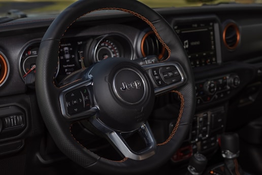 A close-up of a car interior focuses on the steering wheel, which features the Jeep logo. The dashboard includes various controls and a digital display, surrounded by vents with orange accents. The design and details suggest a modern and robust vehicle interior.