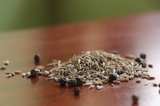 A pile of cumin seeds sits on a wooden surface, interspersed with a few black peppercorns. The background is softly blurred with green tones, highlighting the spices.