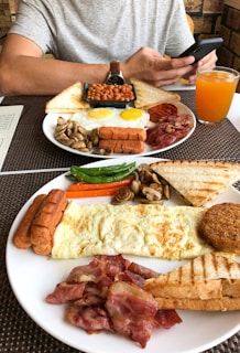 Two plates of a hearty breakfast are arranged on a table. Each plate includes toast, baked beans, sausages, grilled tomatoes, bacon, mushrooms, and eggs. A glass of orange juice and a menu can be seen in the background. A person is sitting at the table, using a smartphone.