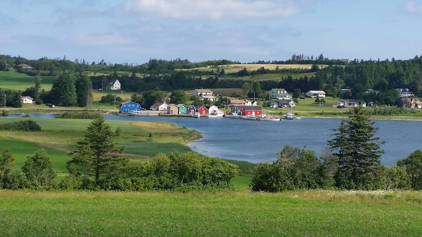 PEI fishing village with coloured boathouses along the harbour, green rolling farmland behind, calm water reflecting the buildings on a summer day