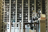 A cozy wine storage room filled with shelves of neatly arranged wine bottles. Wooden tables and chairs are in the foreground, and various wine bottles are placed on the tables. The room has a warm and inviting atmosphere.