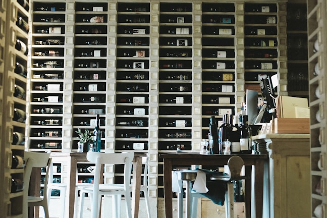 A cozy wine storage room filled with shelves of neatly arranged wine bottles. Wooden tables and chairs are in the foreground, and various wine bottles are placed on the tables. The room has a warm and inviting atmosphere.