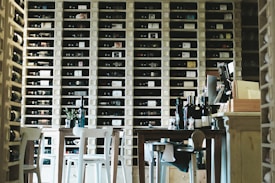 A cozy wine storage room filled with shelves of neatly arranged wine bottles. Wooden tables and chairs are in the foreground, and various wine bottles are placed on the tables. The room has a warm and inviting atmosphere.