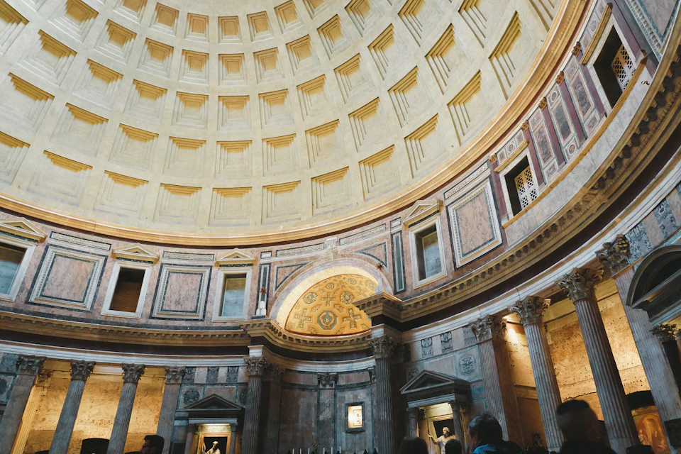 The image depicts a grand, circular room with a high ceiling and intricate designs on its walls. The room is filled with people who appear to be admiring or taking photographs of the architectural marvels around them. The room features numerous columns and arches that add to the overall grandeur of the space. The perspective of the photo suggests it was taken from within the room, providing a unique view of the ceiling and its intricate details.