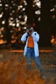 A person in casual attire is standing in a natural setting, holding a camera up to their eye, capturing a photograph. The background features blurred trees, creating a bokeh effect with warm-toned lighting.