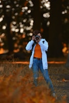 A person in casual attire is standing in a natural setting, holding a camera up to their eye, capturing a photograph. The background features blurred trees, creating a bokeh effect with warm-toned lighting.
