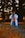 A person in casual attire is standing in a natural setting, holding a camera up to their eye, capturing a photograph. The background features blurred trees, creating a bokeh effect with warm-toned lighting.