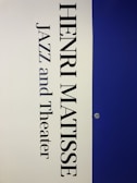 A vertical poster features the name 'Henri Matisse' prominently in large, bold black letters. Below the name, the words 'Jazz and Theater' appear in a similar font style. The background is divided vertically; the left half is a light cream color while the right half is a deep blue. A small metallic fixture is visible on the right side, set against the blue background.