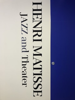 A vertical poster features the name 'Henri Matisse' prominently in large, bold black letters. Below the name, the words 'Jazz and Theater' appear in a similar font style. The background is divided vertically; the left half is a light cream color while the right half is a deep blue. A small metallic fixture is visible on the right side, set against the blue background.