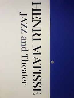 A vertical poster features the name 'Henri Matisse' prominently in large, bold black letters. Below the name, the words 'Jazz and Theater' appear in a similar font style. The background is divided vertically; the left half is a light cream color while the right half is a deep blue. A small metallic fixture is visible on the right side, set against the blue background.