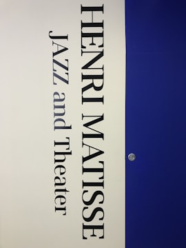 A vertical poster features the name 'Henri Matisse' prominently in large, bold black letters. Below the name, the words 'Jazz and Theater' appear in a similar font style. The background is divided vertically; the left half is a light cream color while the right half is a deep blue. A small metallic fixture is visible on the right side, set against the blue background.