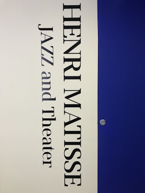 A vertical poster features the name 'Henri Matisse' prominently in large, bold black letters. Below the name, the words 'Jazz and Theater' appear in a similar font style. The background is divided vertically; the left half is a light cream color while the right half is a deep blue. A small metallic fixture is visible on the right side, set against the blue background.