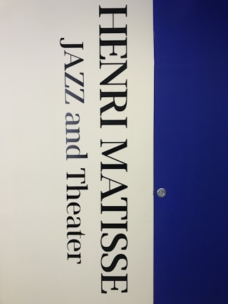 A vertical poster features the name 'Henri Matisse' prominently in large, bold black letters. Below the name, the words 'Jazz and Theater' appear in a similar font style. The background is divided vertically; the left half is a light cream color while the right half is a deep blue. A small metallic fixture is visible on the right side, set against the blue background.