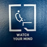 A minimalist pictogram of a person about to fall off an edge is enclosed in a square. Below the image, the words 'WATCH YOUR MIND' are printed in capital letters on a textured blue background.
