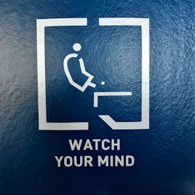 A minimalist pictogram of a person about to fall off an edge is enclosed in a square. Below the image, the words 'WATCH YOUR MIND' are printed in capital letters on a textured blue background.