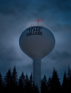 A large, spherical water tower with the text 'Central Texas College' is prominently featured. It stands tall among a silhouetted line of trees against a dark, cloudy sky. A small red light is visible on top of the tower, adding a hint of color to the dim atmosphere.