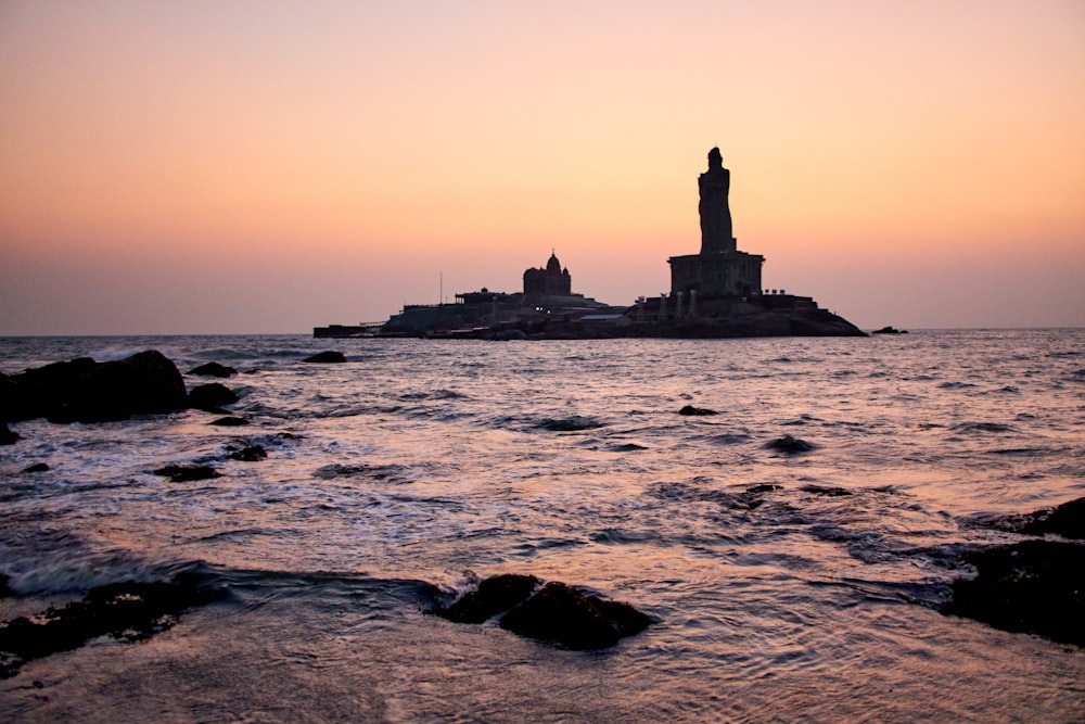 silhouette of lighthouse on rock formation near body of water during sunset