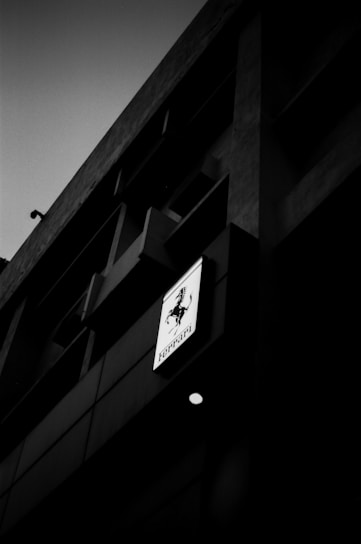 A dark, moody photograph of a building facade, featuring a Ferrari logo on a sign. The image is monochromatic, emphasizing the shadows and architectural lines.